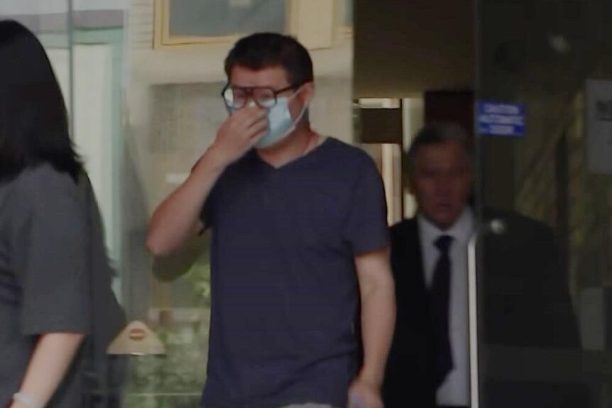 A man wearing glasses and a face mask covers his face as he exits court behind his lawyer.