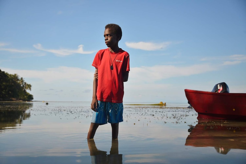 A boy wearing a red t-shirt stands in water above his ankles and looks off to the distance.