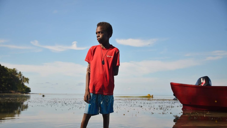 A boy wearing a red t-shirt stands in water above his ankles and looks off to the distance.
