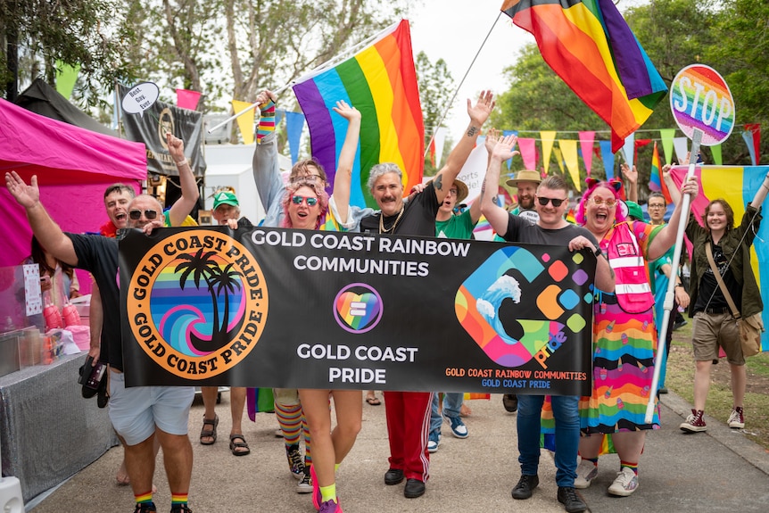 People in colourful clothes wave rainbow flags as they march down a street holding a banner.