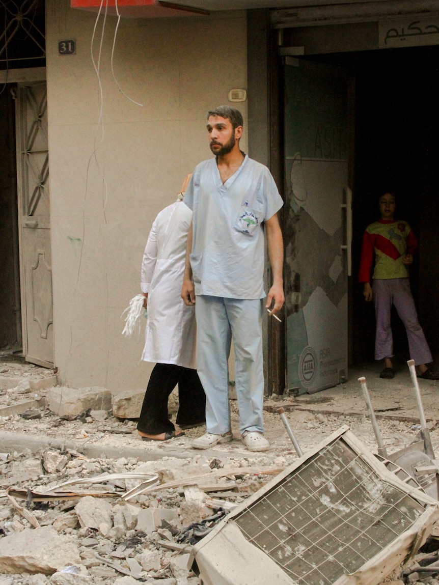 You view a man in dusty, light blue medical scrubs as looks out onto debris strewn across a street from a bombing.