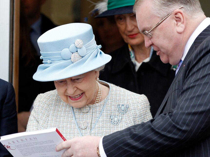 Queen Elizabeth, wearing a blue hat, looks at a book being handed to her by a middle-aged man in pinstripe suit