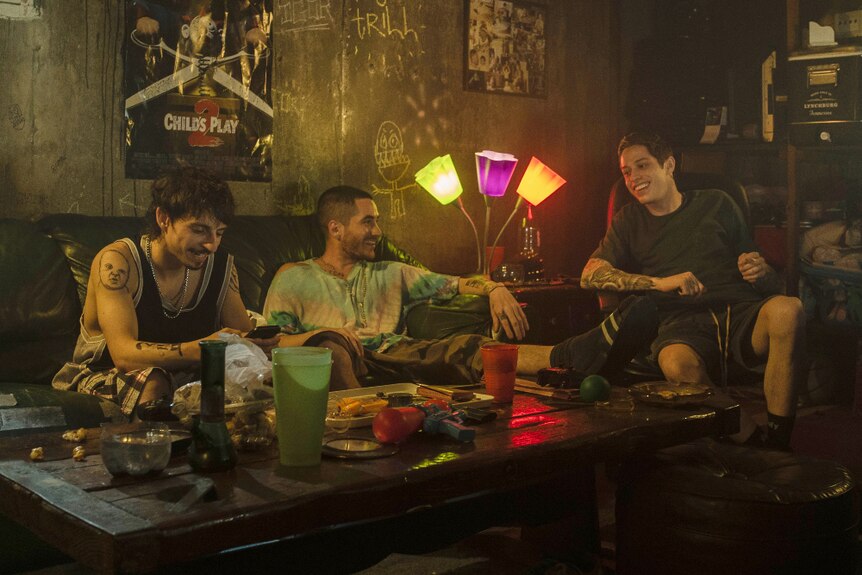 Three tattooed young men sit in dank dark room with movie posters and chalk graffiti on wall and table full of plates and cups.