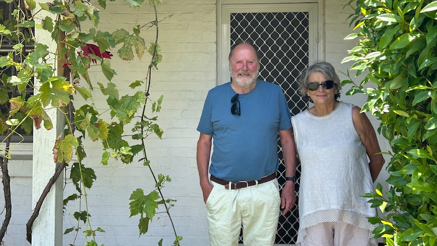 A man and woman stand in front of a white cottage with vines hanging down.