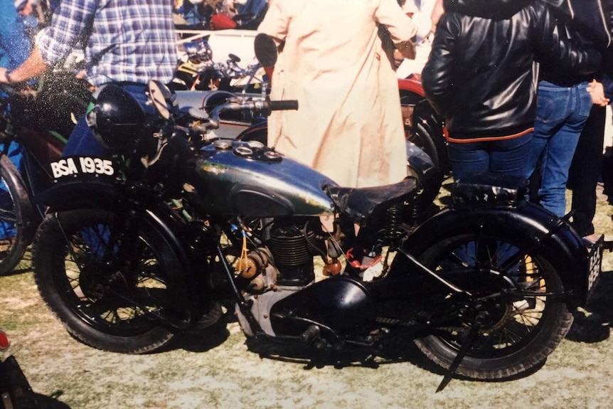 An historic motorcycle is surrounded by people and other vehicles on a lawn