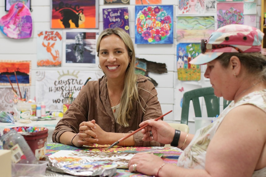Woman smiling at the camera with art in the background, and woman next to her painting a picture on the table.
