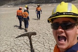 Tom Maslin, mining engineering graduate, smiling holding prospectors pic with unidentifiable workers in hi-vis in background