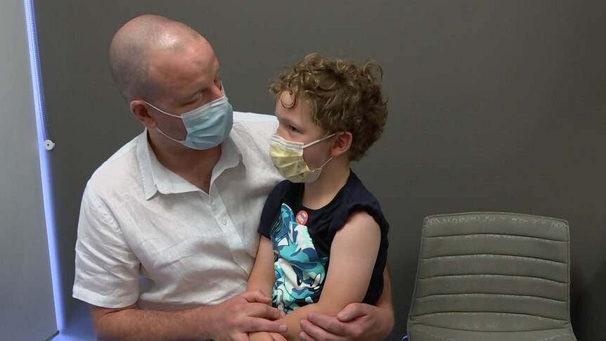 A boy sitting on his dad's knee before getting his COVID vaccination with his sleeve rolled up, they both wear masks
