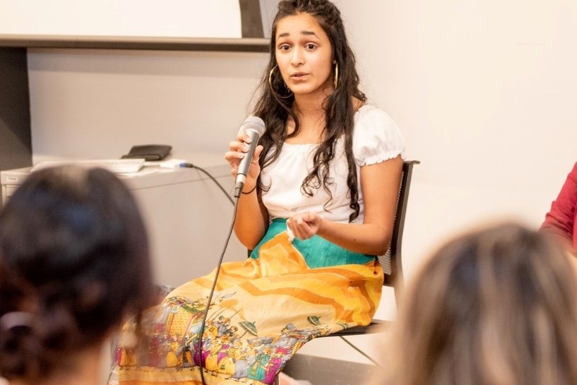 Yatha Jain speaking at an event, pictured with a microphone in her hand.