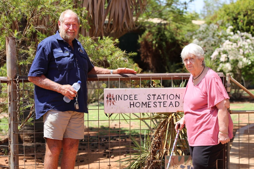 An older man and woman standing by a gate with a sign that says 'Indee Station Homestead'