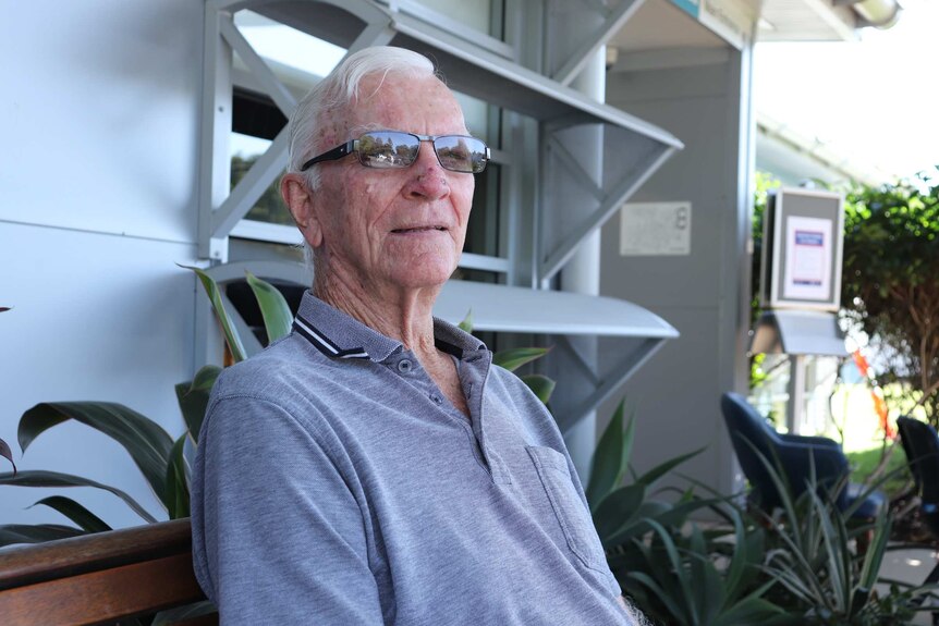 An elderly man sits in front of what appears to be a hospital clinic. He is wearing what appear to be transition lenses.