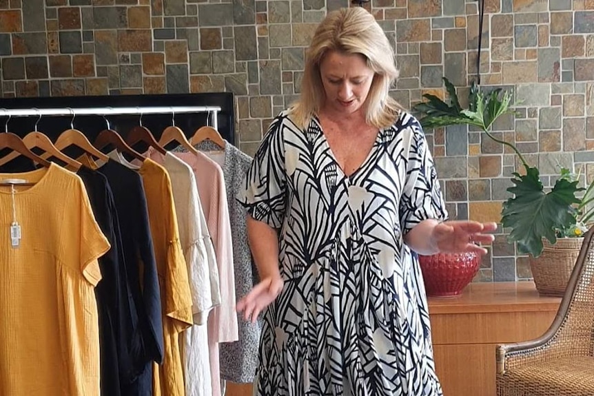 woman with blonde hair in white flowy dress with black patterns next to clothes rack of tops and dresses