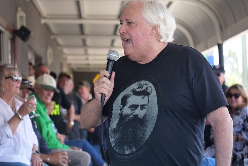 Clive Palmer wearing a Ned Kelly shirt, holds a microphone mid speech, punters in the background.