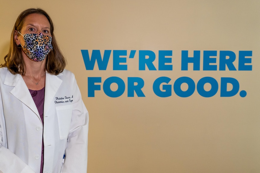 A female doctor wearing a printed face mask and lab coat standing in front of a wall with text.