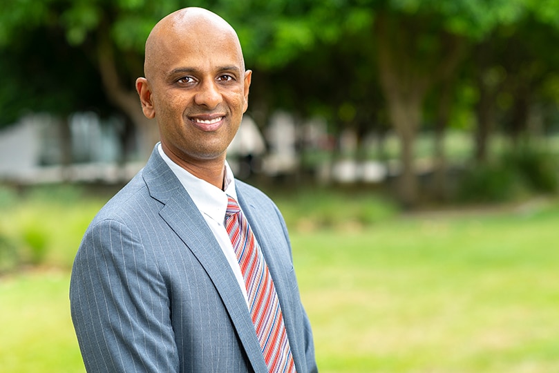 A slim man in a sharp suit stands outside smiling.