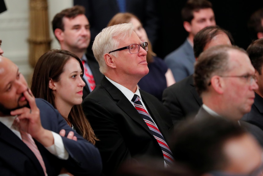 Jim Hofts in a suit sits among a group of people.