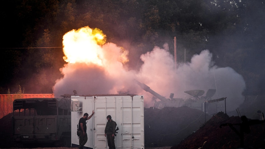 Two soldiers stand by a white shipping container as a large vehicle fires in a flash of smoke and flame behind them.