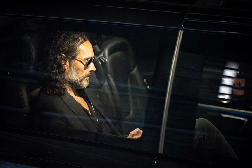 Russel Brand sits on the backseat of a car wearing sunglasses
