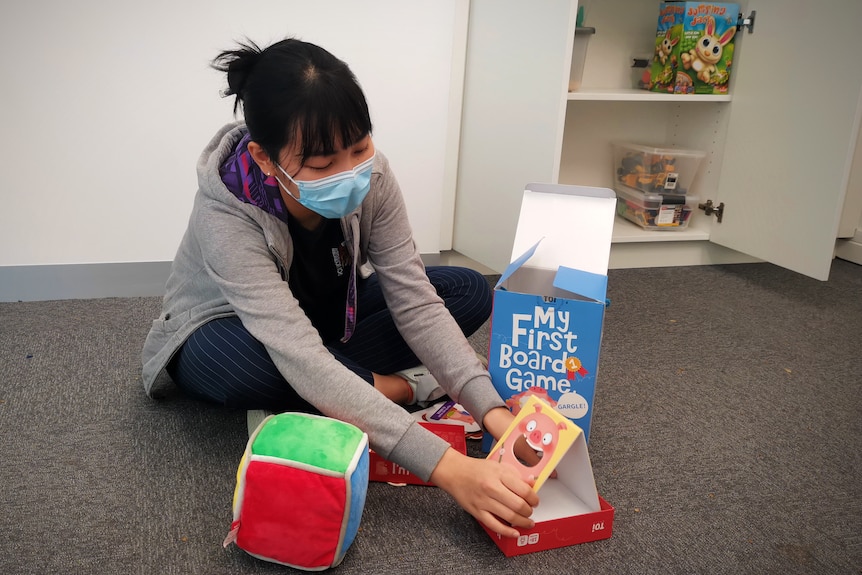 A young woman wearing a mask sits on the floor with a box open that says 'my first board game'