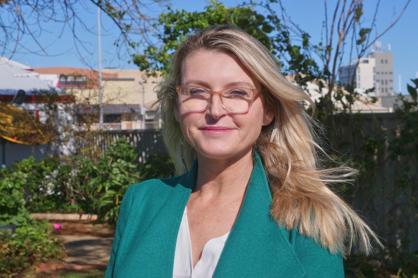 Headshot of a blonde-haired woman wearing glasses, white shirt and green jacket. 