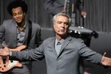David Byrne in grey suit stands spot-lit on stage with arms stretched wide, and barefoot musicians in grey suits in background.