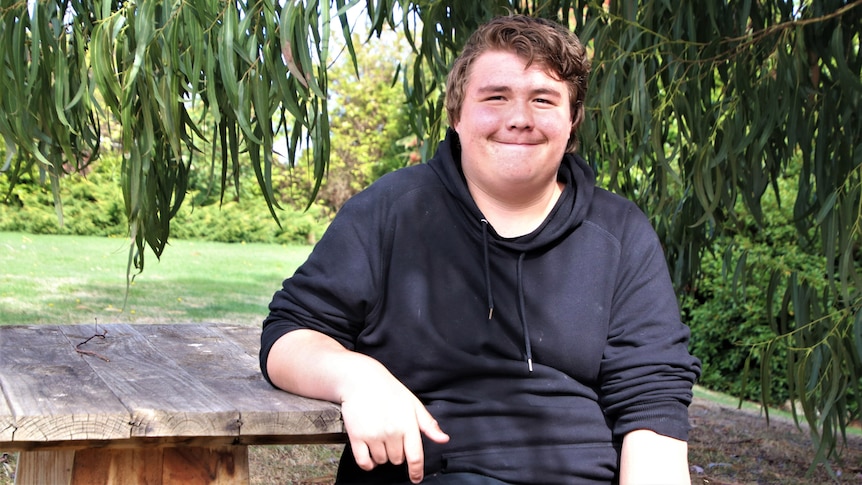A teenage boy sits at a park bench in a dark hoody, smiling 