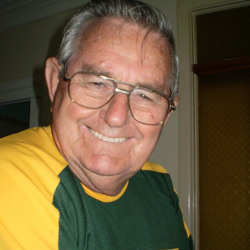 Geoffrey Meyers, wearing a blue-and-gold Tshirt and gold-rimmed glasses, smiles widely at the camera.