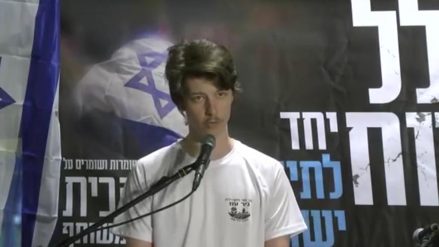 A picture of a young man standing at a lectern giving a speech with Israeli flags behind him.