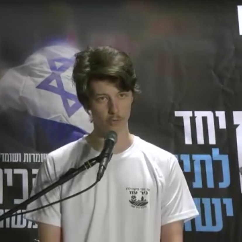 A picture of a young man standing at a lectern giving a speech with Israeli flags behind him.