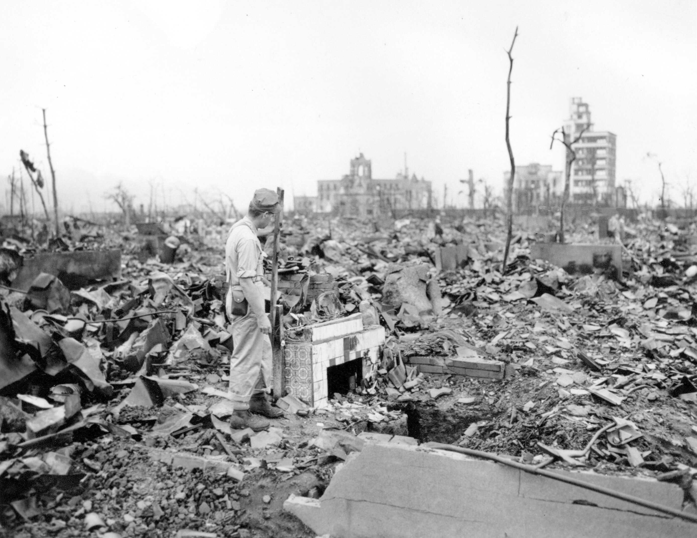 A man wearing a cap standing amid mounds of destroyed rubble and a tiled fireplace, next to trees stripped of their leaves