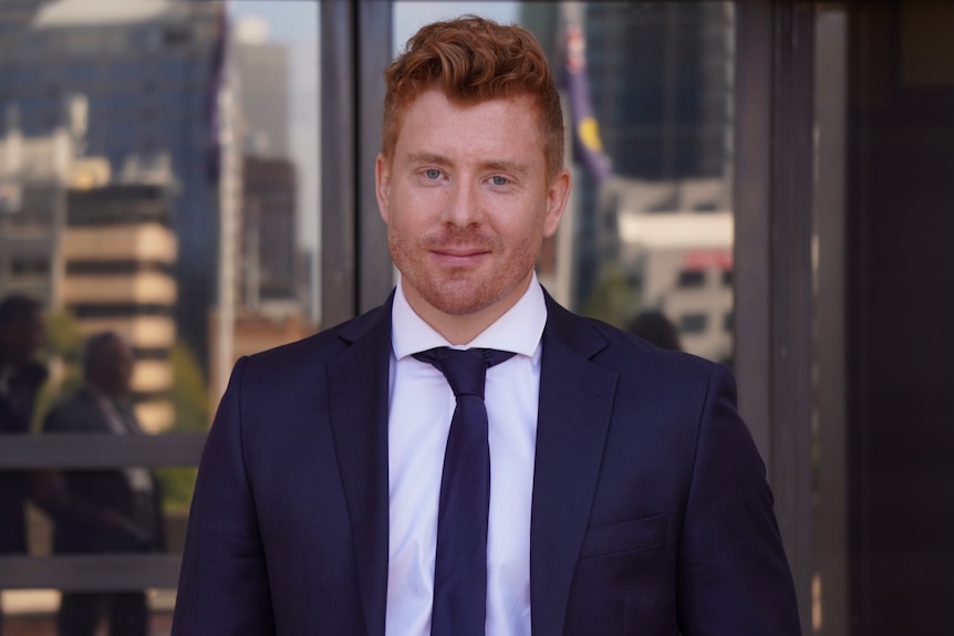 Wilson wears a navy blue suit and tie, looking into the camera on the steps of WA's Parliament House.