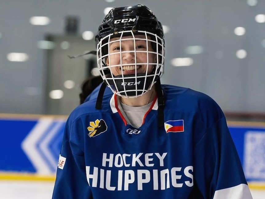 一名女冰球运动员身穿印有“菲律宾冰球”字样的球衣，头戴面罩头盔，对着镜头微笑。
