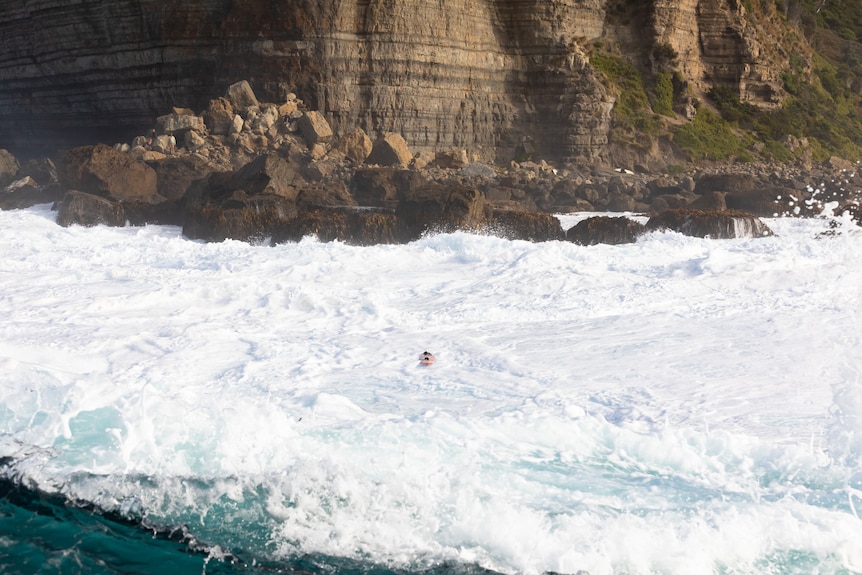 A surfboard can be seen in churning white water near a rocky coastline and cliffs.