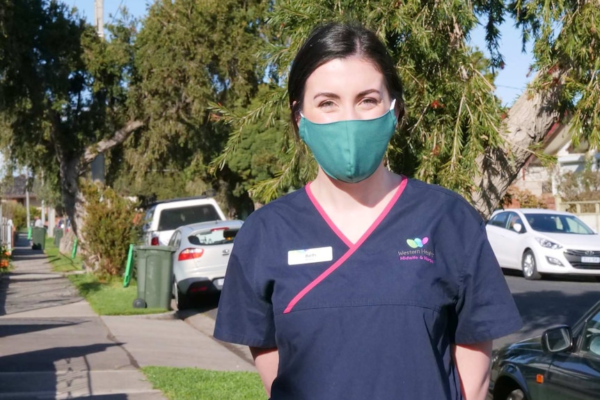 A woman standing in blue hospital scrubs with a green mask on.