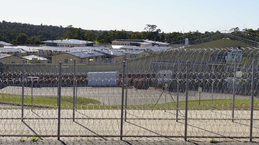 Wide shot of buildings in Acacia prison, with barbed wire fences in the foreground and bushland behind the prison.