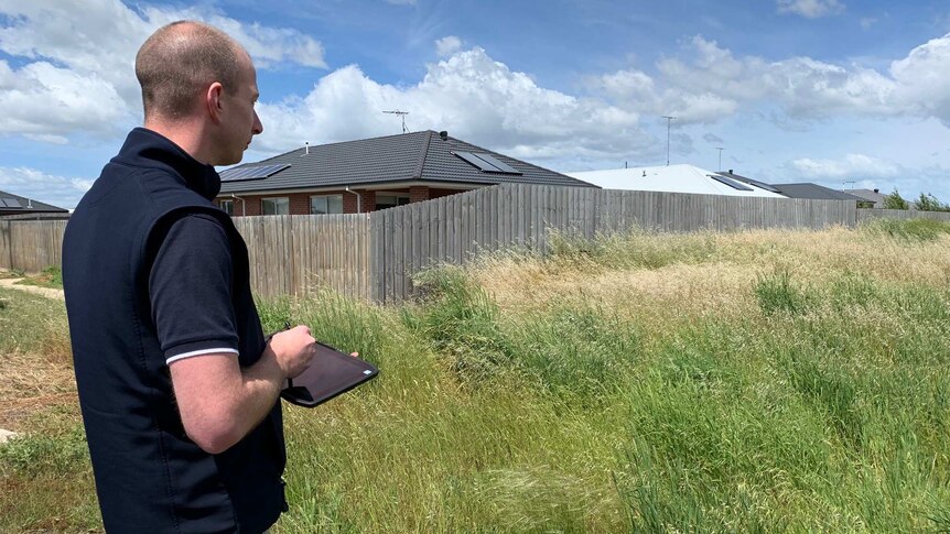 A man with an iPad stands by a field of long green grass which borders residential homes.