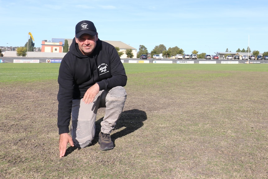 New Port Lincoln Curator Hayden Parker dressed in a black hoodie and black hat bending down on to touch turf pitch