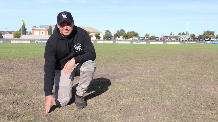 New Port Lincoln Curator Hayden Parker dressed in a black hoodie and black hat bending down on to touch turf pitch