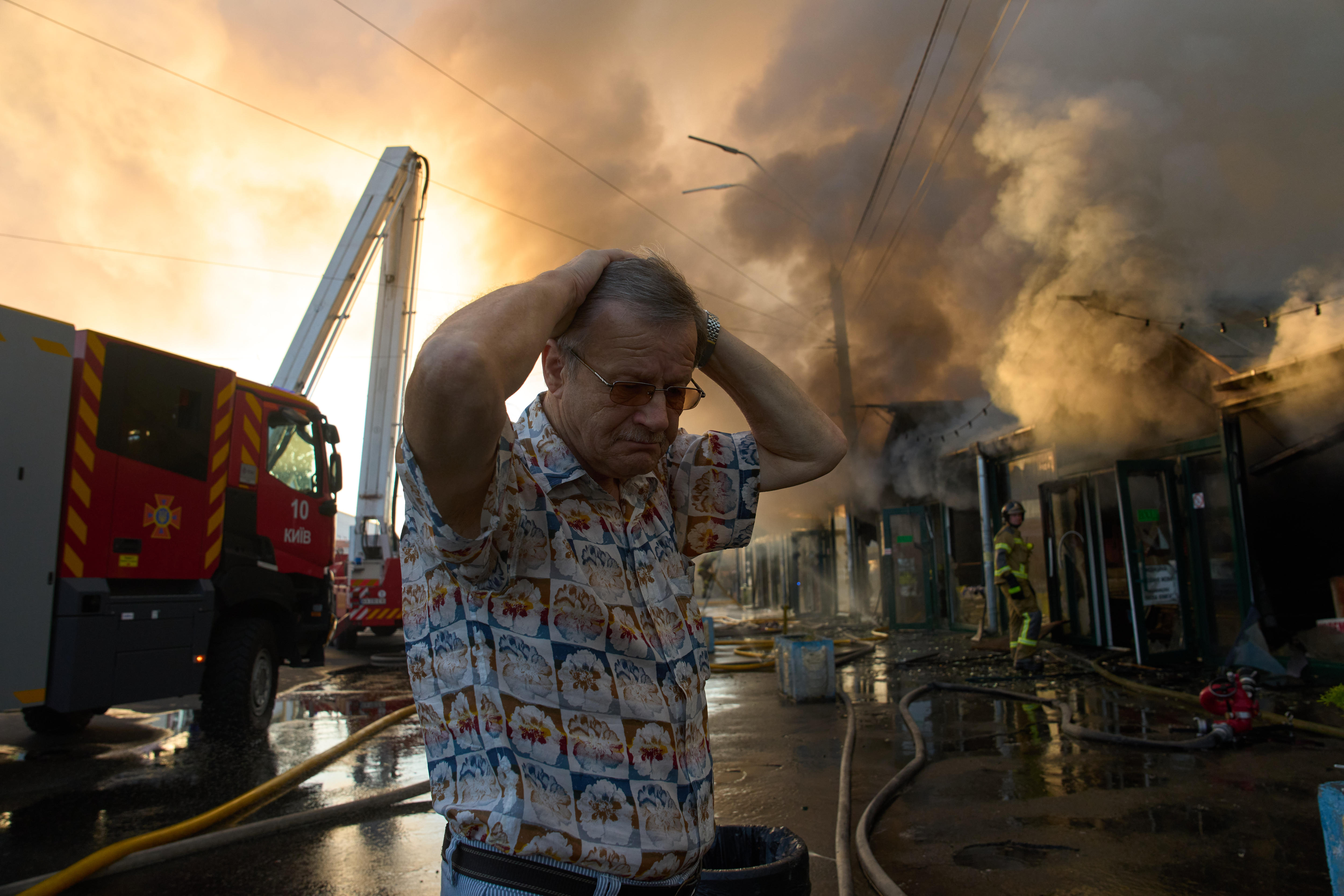 An older man with his hands on his head looking dismayed as smoke rises from a building behind him.
