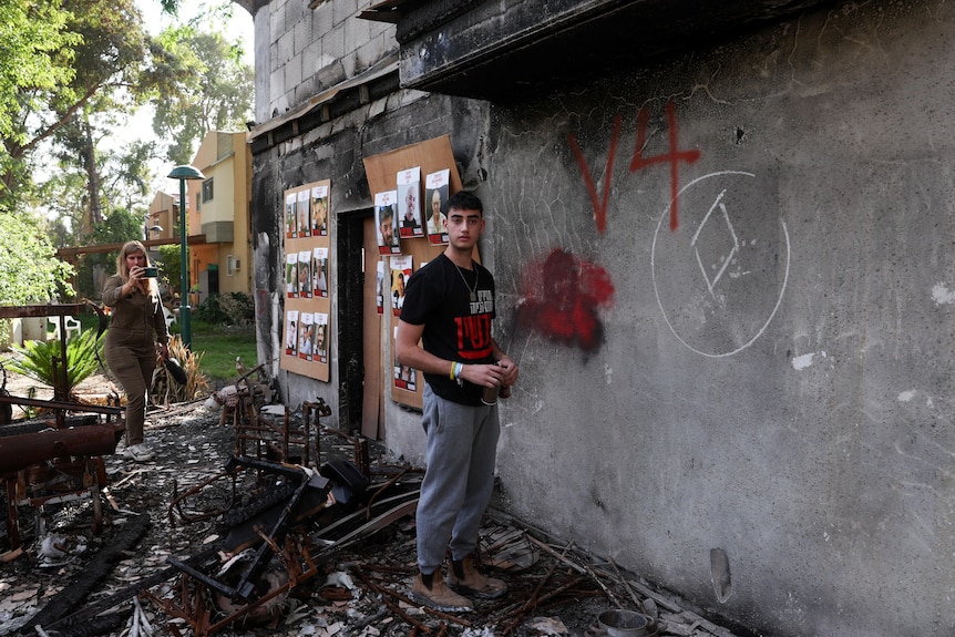 A young man in black Tshirt stands outside a concrete structure with spray paint and photos tacked onto it, debris on the ground