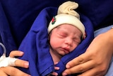 A newborn infant wrapped in bedding and wearing a beanie is held by her mother in a hospital bed.