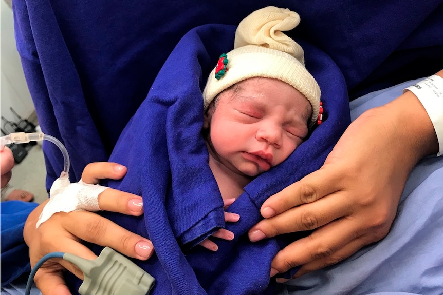 A newborn infant wrapped in bedding and wearing a beanie is held by her mother in a hospital bed.