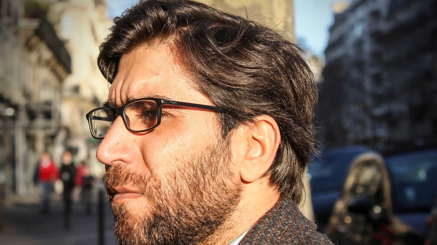 Omar wearing a suit jacket, collared shirt with a clipped beard and glasses sits on a bench.