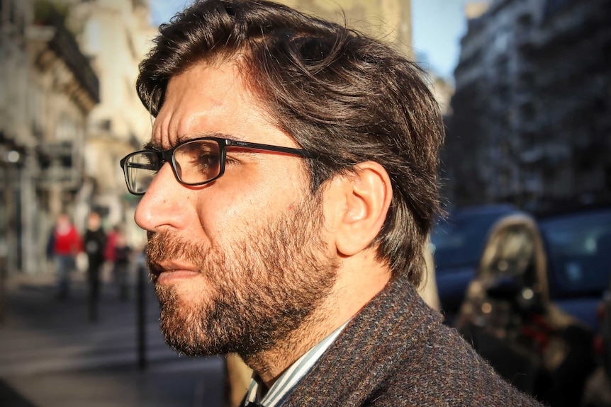 Omar wearing a suit jacket, collared shirt with a clipped beard and glasses sits on a bench.