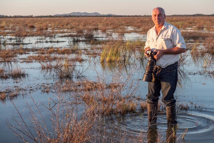 An older white man standing in a swamp with a large camera