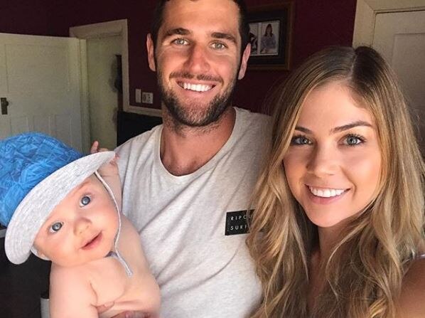 Jack holds his baby and poses with his wife, smiling at the camera