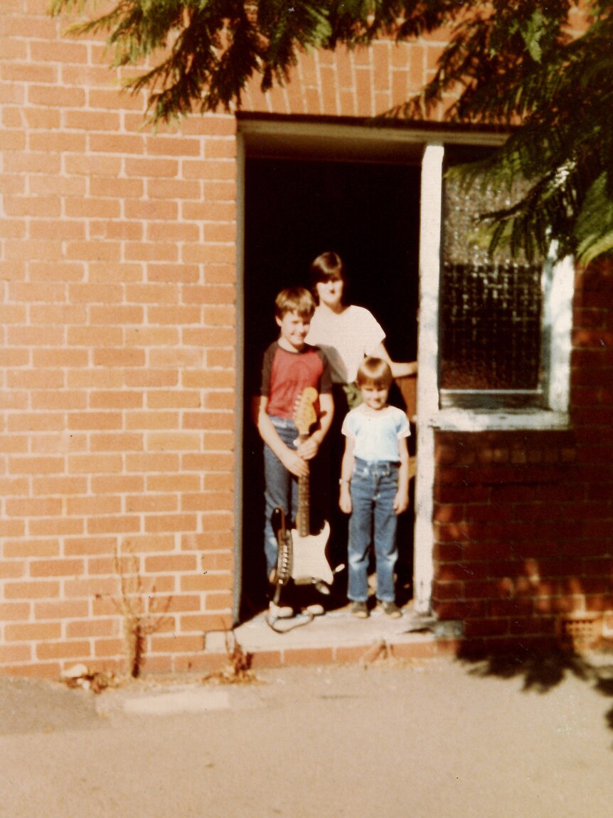 A boy stands in the doorway of a house carrying a guitar, with a woman and a younger boy next to him. 