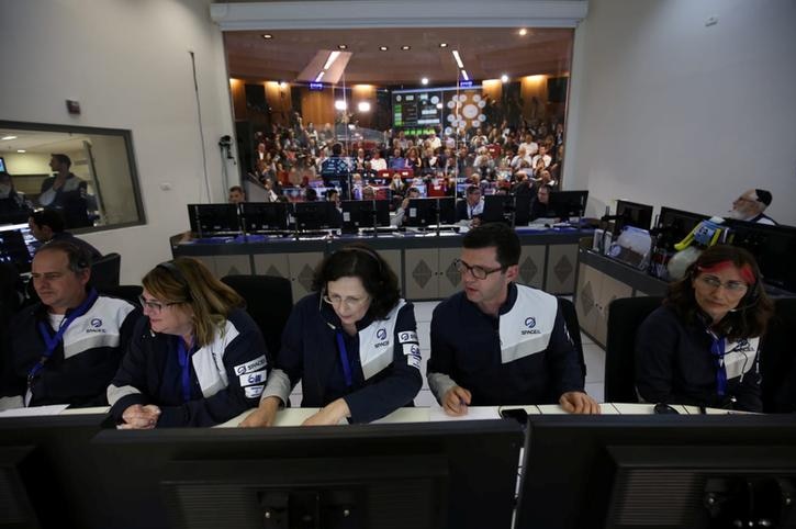 Five control room workers are seen in a room with a glass wall behind them and a room full of people sat beyond it.