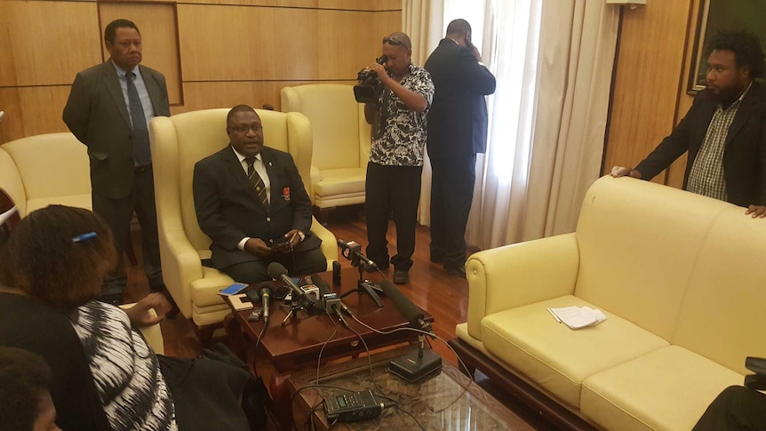 Papua New Guinea's electoral commissioner sitting on a yellow armchair in a meeting room with several onlookers and microphones