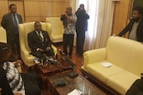 Papua New Guinea's electoral commissioner sitting on a yellow armchair in a meeting room with several onlookers and microphones